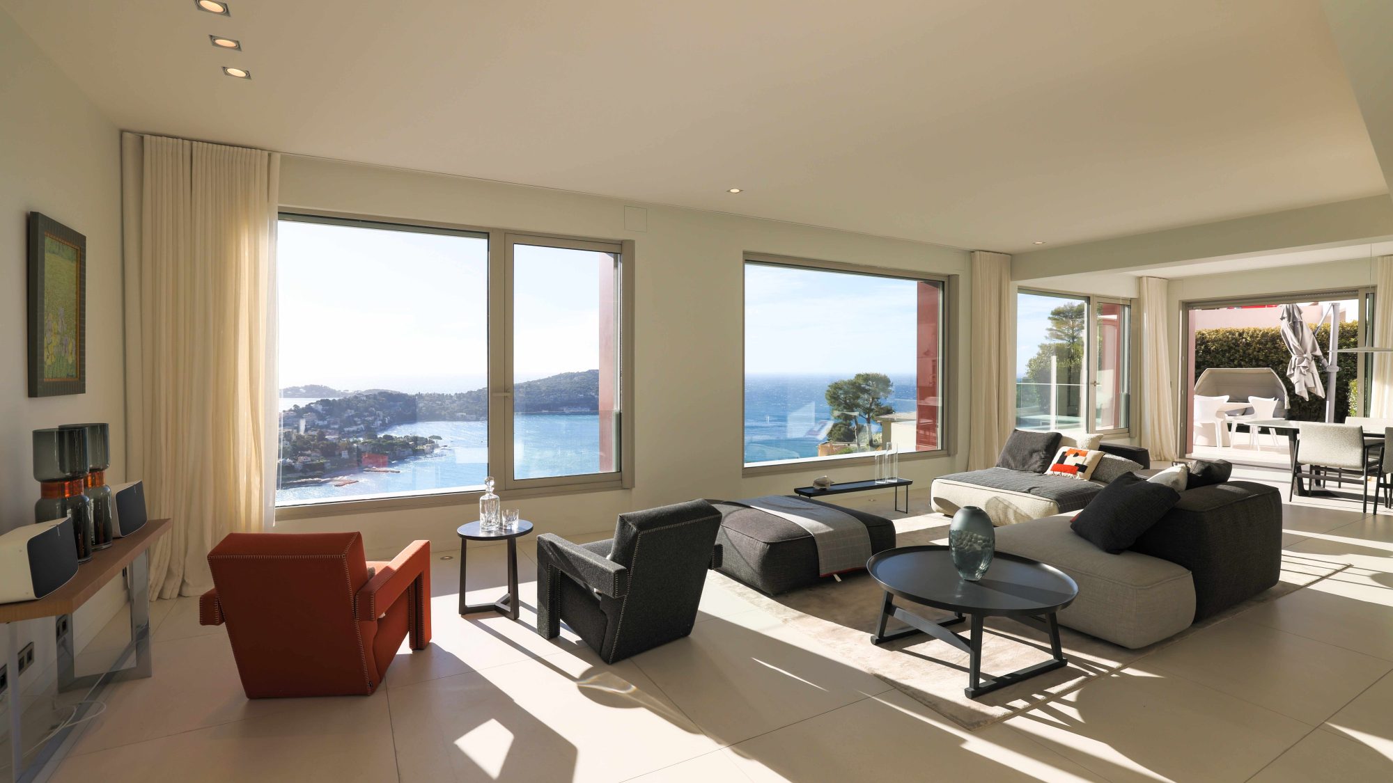 Interior of a luxury villa rental from Vista Magnifika. In a large, bright, modern living room, armchairs and a sofa face the Mediterranean Sea.  